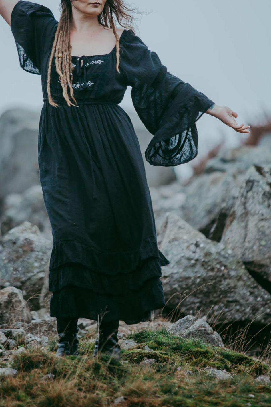 A person standing in a natural black dress with embroidered details, a lace chest panel, and a tie-up front, on a grassy area with rocks in the background.