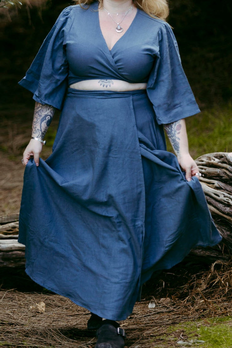 Woman in a blue dress standing in a natural setting with trees and logs.