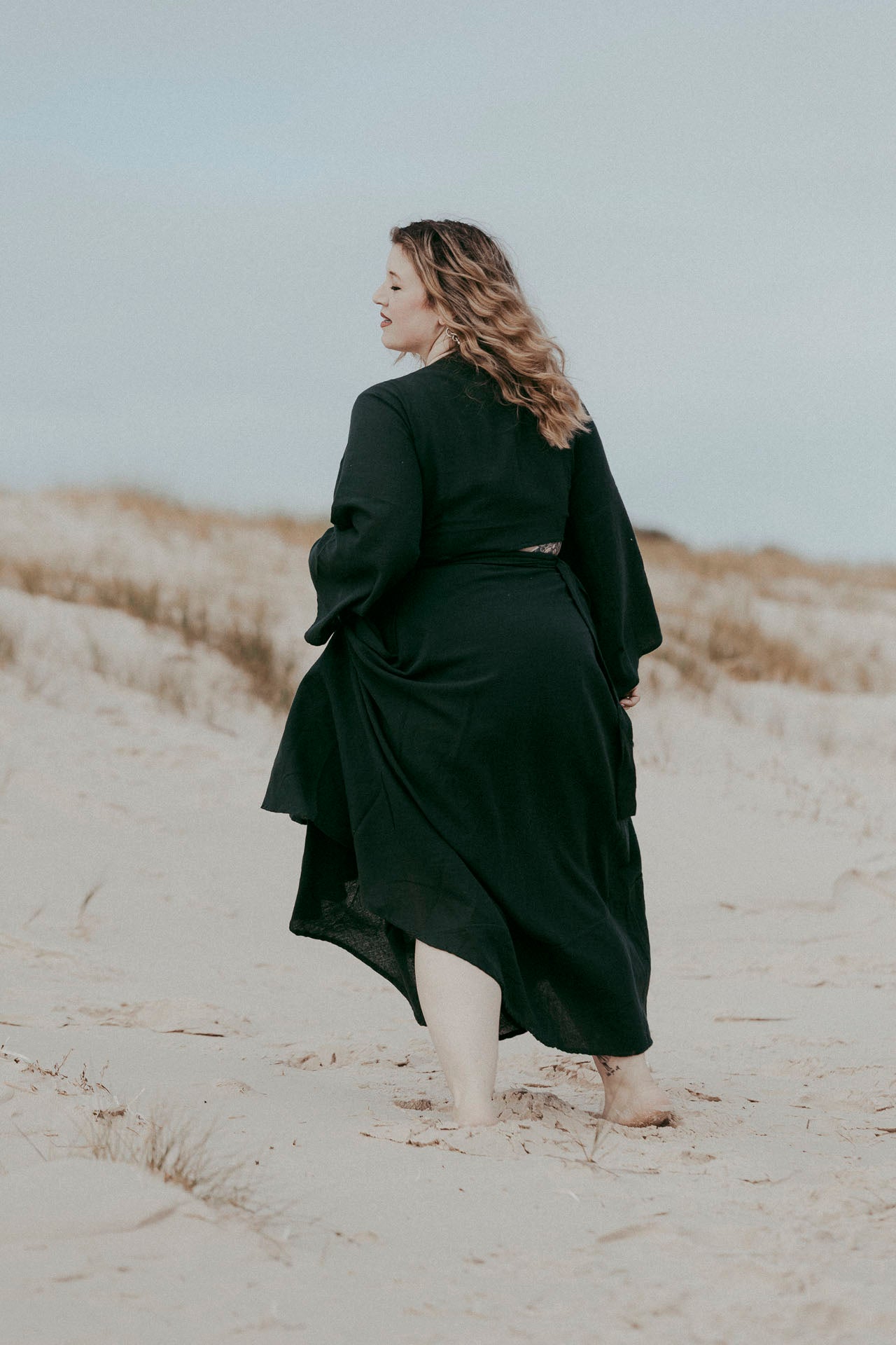 Woman in a long dark green coat standing on a sandy beach with a blurred background