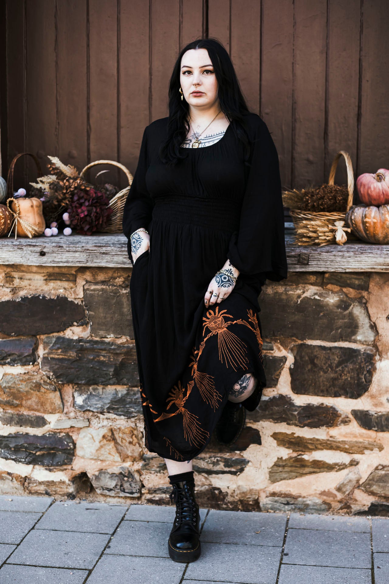 Person wearing a black dress with embroidery standing in front of a stone wall with pumpkins and decorations.
