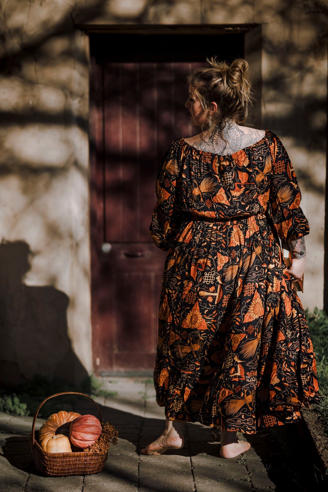 Woman in a patterned dress standing in front of a stone wall with a basket of bread and pumpkins.