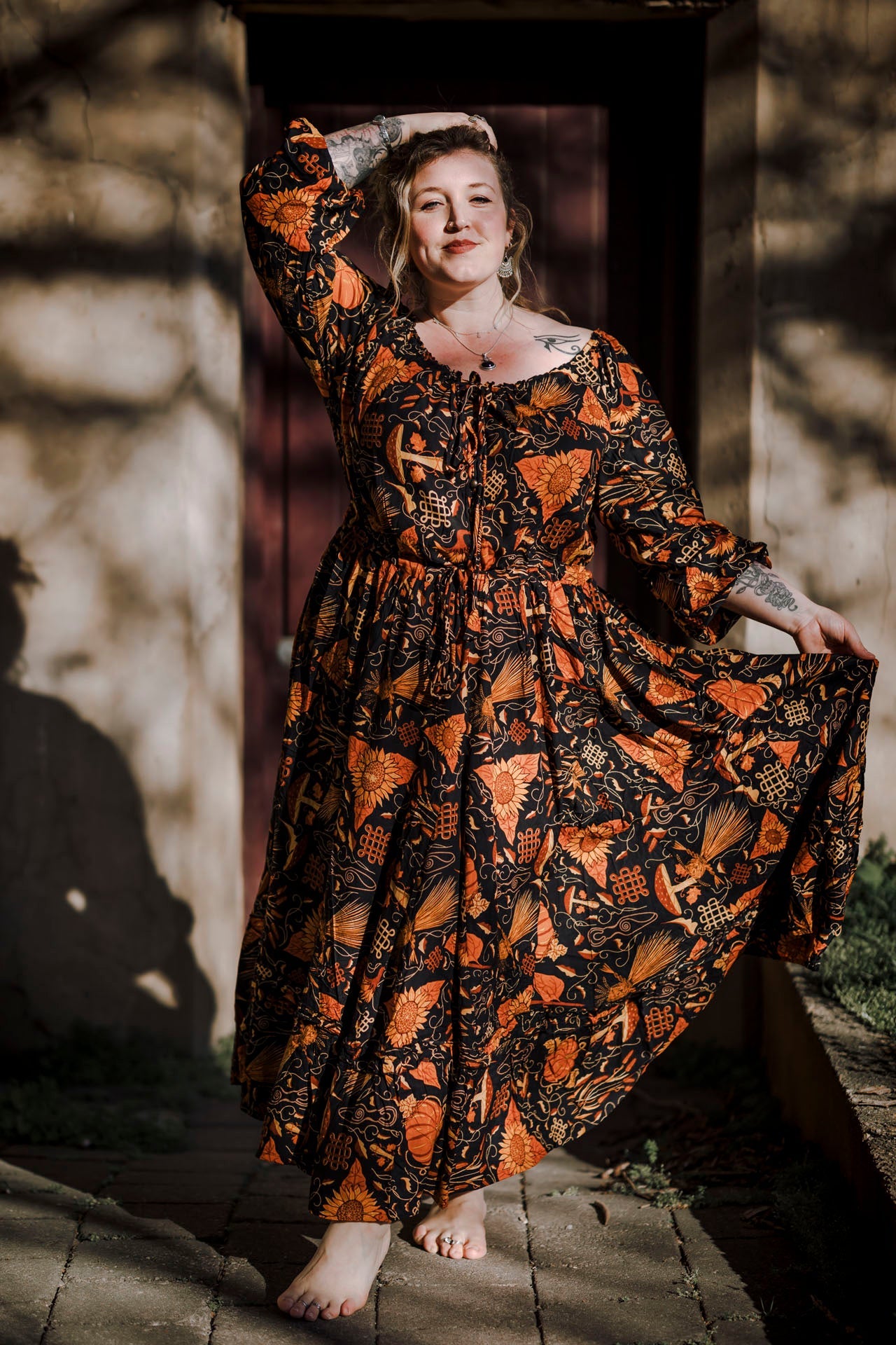 Woman in a long, patterned dress standing outdoors with a stone wall and wooden door in the background.