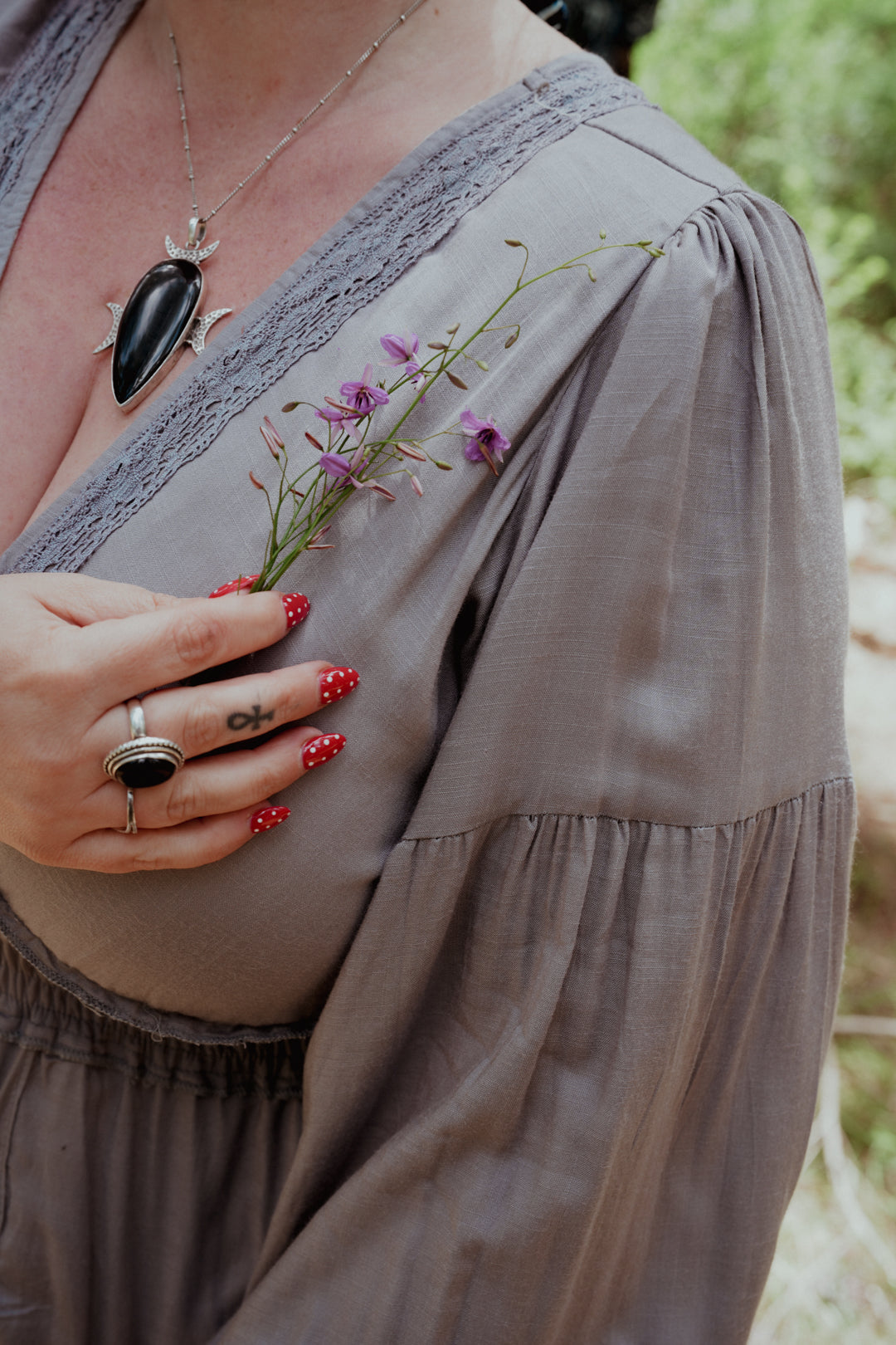 Person wearing a necklace with a large black stone, holding small purple flowers, with a blurred natural background.